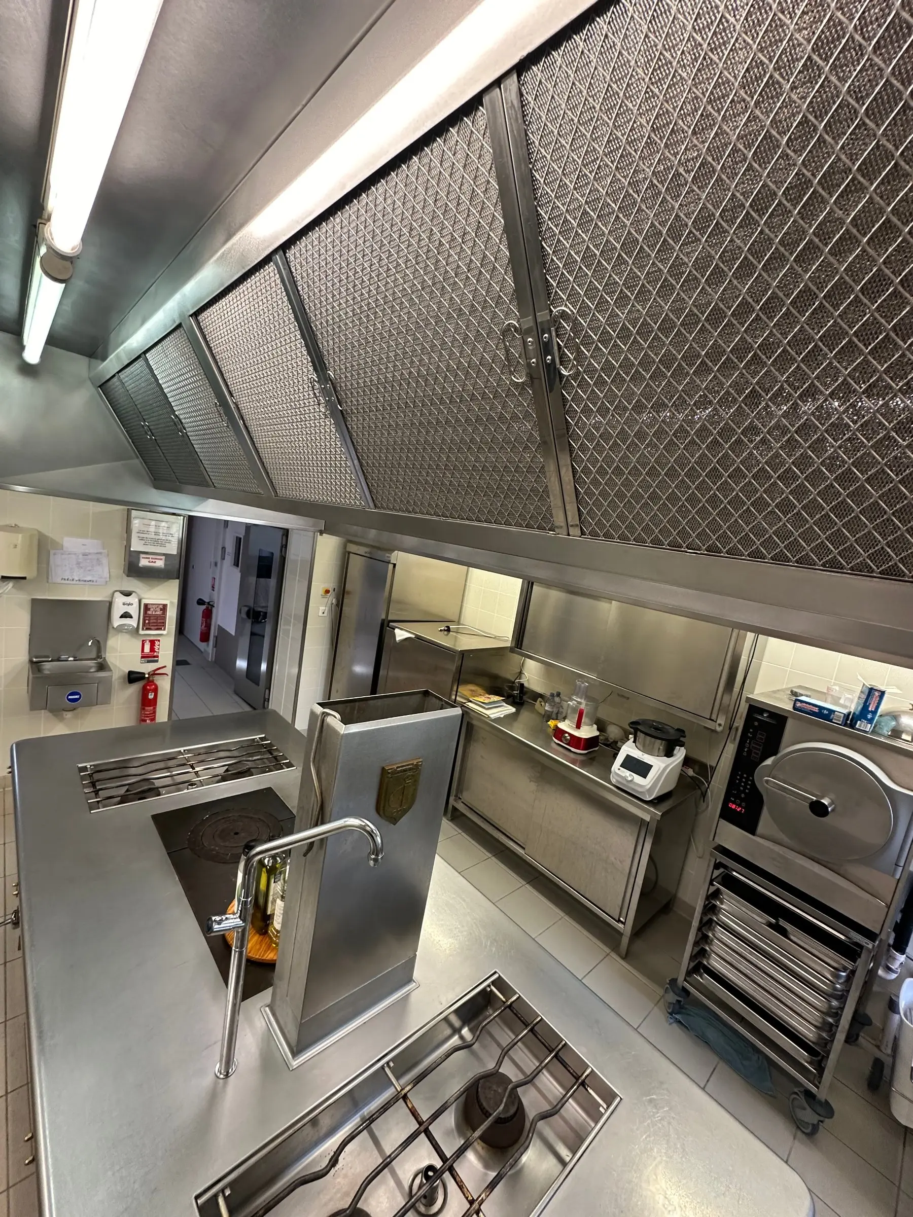 Plan de travail de cuisine professionnelle en inox avec hotte et filtres métalliques; espace de travail équipé, éviers, et matériel de cuisson. Plan de travail de cuisine professionnelle en inox avec hotte et filtres métalliques; espace de travail équipé, éviers, et matériel de cuisson.